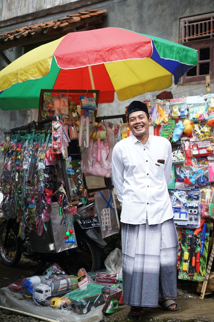 Street vendor smiling in front of a colorful toy stall under a rainbow umbrella on a vibrant day.